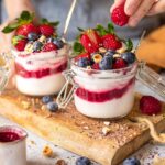Das Foto zeigt, wie eine Hand vorsichtig eine Himbeere zu einem Glas mit geschichtetem Joghurt und Beerenkompott hinzufügt, garniert mit "Bio Porridge Food Finisher" und frischen Früchten nach Rezept.
