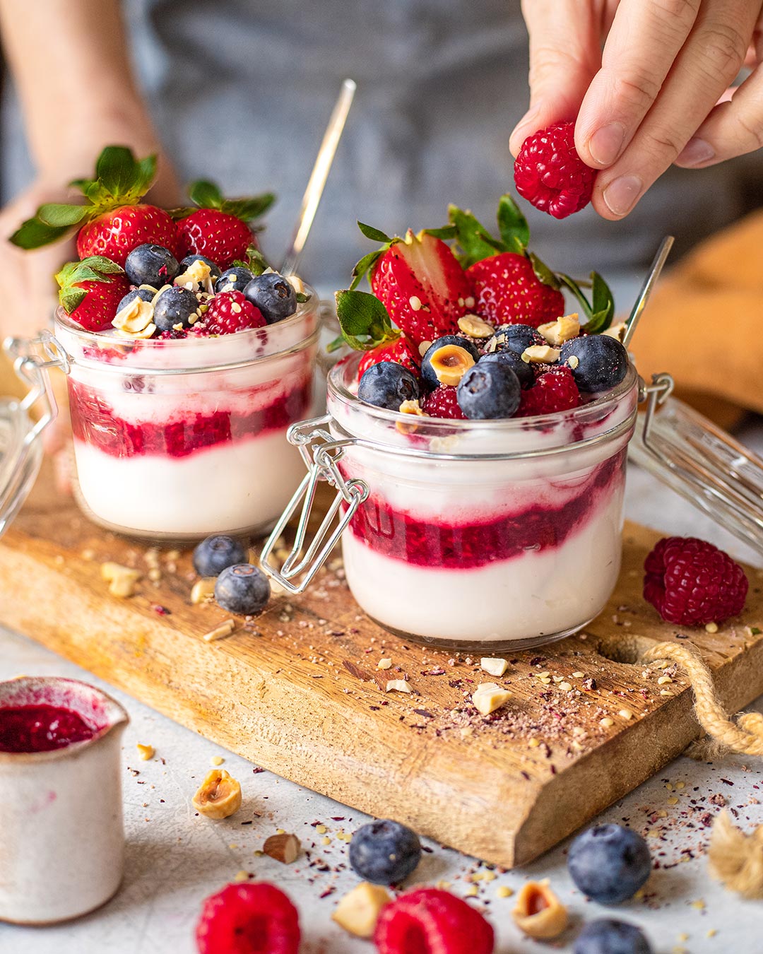 Das Foto zeigt, wie eine Hand vorsichtig eine Himbeere zu einem Glas mit geschichtetem Joghurt und Beerenkompott hinzufügt, garniert mit "Bio Porridge Food Finisher" und frischen Früchten nach Rezept.