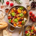 Das Foto zeigt einen mediterranen Pasta-Salat, verfeinert mit "Food Finisher Mediterran", neben frischen Zutaten und einem Stück Brot nach Rezept.