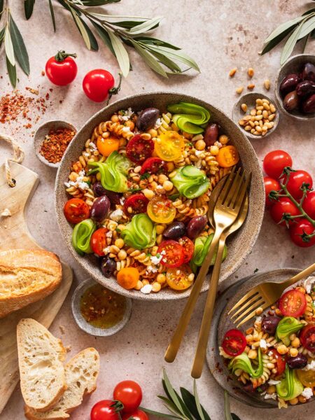 Das Foto zeigt einen mediterranen Pasta-Salat, verfeinert mit "Food Finisher Mediterran", neben frischen Zutaten und einem Stück Brot nach Rezept.