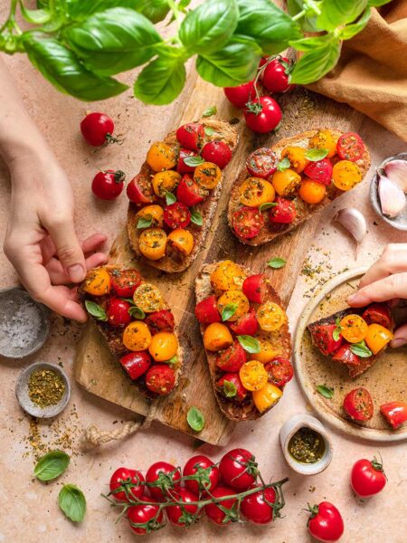 Das Foto zeigt rustikale Tomatenbruschettas, verfeinert mit "Food Finisher Tomaten Zauber" und frischem Basilikum, angerichtet auf einem Holzbrett nach Rezept.