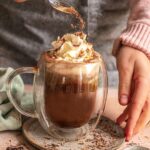 Das Foto zeigt heiße Kokosnuss-Schokolade nach Rezept, garniert mit Schlagsahne und Kakao, serviert in einer klaren Tasse.