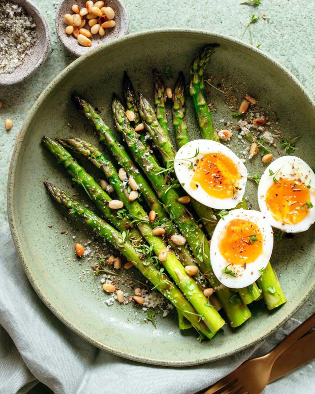 Das Foto zeigt frisch zubereiteten grünen Spargel mit halbierten, weichgekochten Eiern, bestreut mit Pinienkernen, Food Finisher und Parmesan nach Rezept.