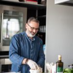 Spitzenkoch Luis Dias aus Köln, in einer Küche, wie er mit Lebensmitteln arbeitet. Auf dem Tisch liegen übriggebliebene Zutaten wie Brot, Gemüse und Kräuter, die er kreativ zu einem neuen Gericht kombiniert. Luis gibt Tipps zur Vermeidung von Lebensmittelverschwendung.