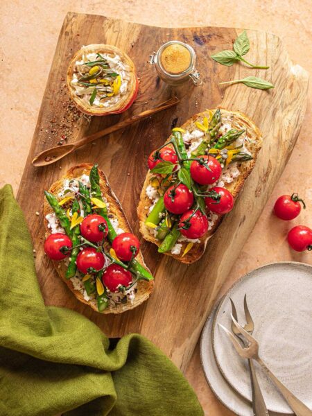 Das Foto zeigt zeigt köstliches Sauerteigtoast mit Ricotta, gegrilltem Spargel und Kirschtomaten nach Rezept, verfeinert mit BIO BRATEN FOOD FINISHER, präsentiert auf einem rustikalen Holzbrett.