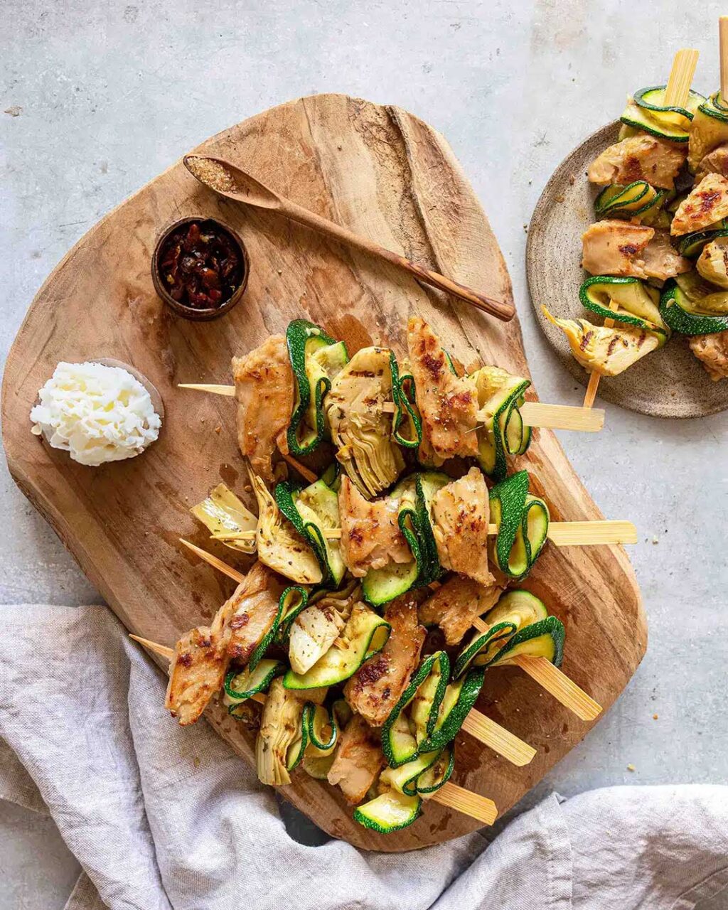Vier Zitronen-Hähnchenspieße, kombiniert mit Artischocke und Zucchini, angerichtet auf einem rustikalen Holzbrett. Verfeinert mit dem Bio Braten Finisher von Luis Dias, der mit seiner Salz-Pfeffer-Mischung dem Gericht den letzten Schliff verleiht.