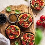 Mini Tomaten-Tarten auf einem Holzbrett, belegt mit Kirschtomaten und Basilikumpesto, bestreut mit Bio Tomate Food Finisher von Luis Dias, garniert mit frischem Basilikum.