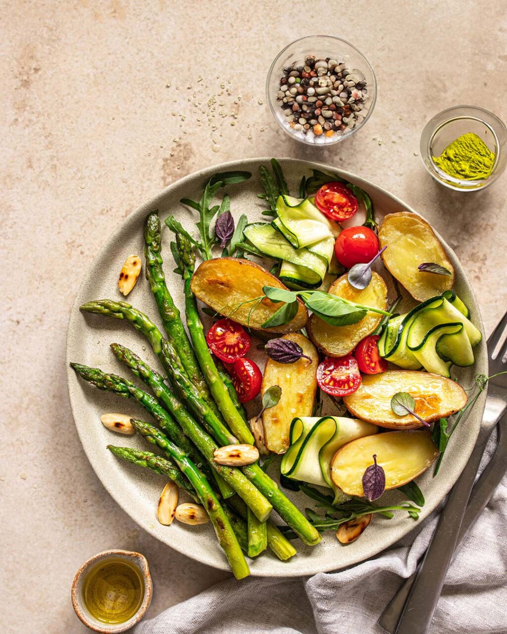 Spargelsalat mit gebackenen Kartoffeln, serviert auf einem hell-grauen Keramikteller. Fertiggestellt mit Kirschtomaten, gerollter Zucchini und Mandelplättchen und verfeinert mit dem Bio Pommes de Terre Food Finisher von Luis Dias.