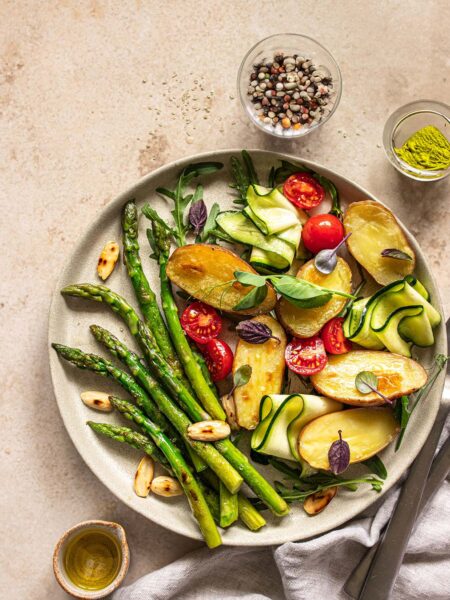 Spargelsalat mit gebackenen Kartoffeln, serviert auf einem hell-grauen Keramikteller. Fertiggestellt mit Kirschtomaten, gerollter Zucchini und Mandelplättchen und verfeinert mit dem Bio Pommes de Terre Food Finisher von Luis Dias.