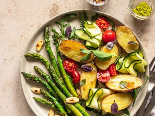 Spargelsalat mit gebackenen Kartoffeln, serviert auf einem hell-grauen Keramikteller. Fertiggestellt mit Kirschtomaten, gerollter Zucchini und Mandelplättchen und verfeinert mit dem Bio Pommes de Terre Food Finisher von Luis Dias.