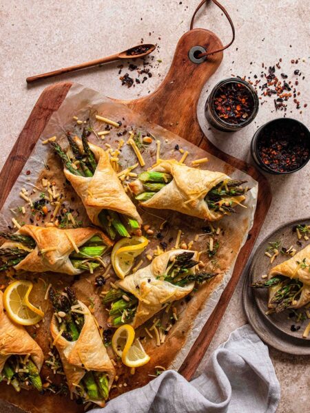 Kleine Spargel im Blätterteig Häppchen präsentiert auf einem Holzbrett. Gefüllt mit veganem Schinken und veganem Käse und verfeinert mit dem Bio Pommes de Terre Food Finisher von Luis Dias.