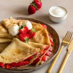 Erdbeer-Crêpes mit einer Füllung aus Erdbeeren, Vanilleschote und den Bio Food Finisher von Luis Dias. Schön angerichtet auf einem grauen Keramikteller mit halbierten Erdbeeren und zwei Kugeln feinem Vanilleeis.