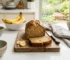 Professionelles Food-Foto: Ein frisch gebackenes, angeschnittenes Bananenbrot auf einem rustikalen Holzbrett in einer hellen, freundlichen Küche. Neben dem Brot liegen Bananenscheiben, im Hintergrund eine Schüssel mit reifen Bananen und Küchenutensilien, natürliches Tageslicht.