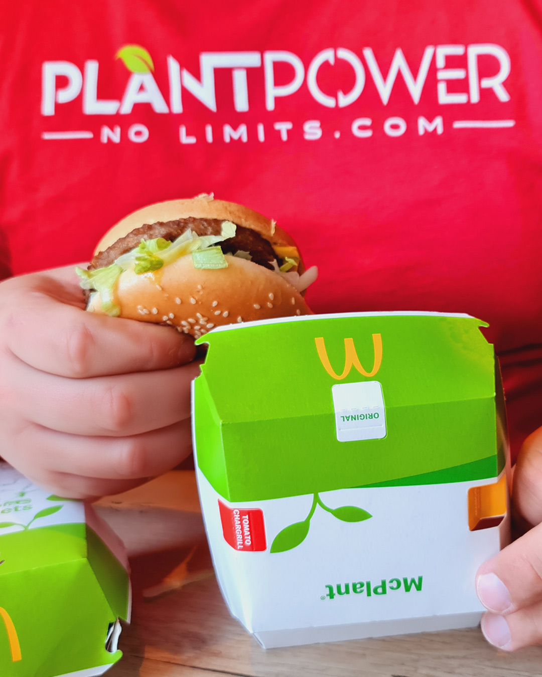 Eine Testperson von PlantPower-NoLimits.com trägt ein rotes T-Shirt mit dem Logo von PlantPower-NoLimits und hält einen McPlant Burger von McDonald's in der rechten Hand. In der linken Hand hält die Person die McPlant-Verpackung. Daneben ist eine teilweise sichtbare McPlant Nuggets-Box zu sehen. Die Verpackung von McDonald's zeigt ein veganes Symbol mit zwei Blättern und der Aufschrift „McPlant“.