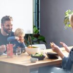 Eine fröhliche Familie sitzt an einem Tisch während ihres Airbnb-Urlaubs. Die Eltern und das Kind lachen und genießen ihr Essen, das mit Bio Food Finisher Gewürzmischungen verfeinert wurde. Die Gewürzmischungen stehen auf dem Tisch, während die Familie die gemeinsame Mahlzeit in entspannter Atmosphäre genießt.