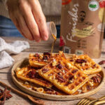 Eine Hand gießt Sirup über knusprige Kürbiswaffeln, die mit Nüssen garniert und mit BIO KAKAO FOOD FINISHER bestäubt sind. Die Waffeln liegen auf einem rustikalen Teller, der auf einem Holztisch angerichtet ist. Im Hintergrund ist die Packung des BIO KAKAO FOOD FINISHER sichtbar.