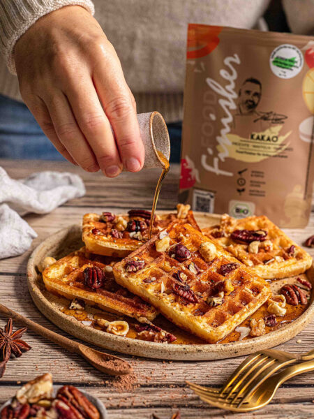 Eine Hand gießt Sirup über knusprige Kürbiswaffeln, die mit Nüssen garniert und mit BIO KAKAO FOOD FINISHER bestäubt sind. Die Waffeln liegen auf einem rustikalen Teller, der auf einem Holztisch angerichtet ist. Im Hintergrund ist die Packung des BIO KAKAO FOOD FINISHER sichtbar.