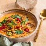 Eine Schüssel mit roter Thai-Kokos-Suppe, garniert mit Koriander, Frühlingszwiebeln und Chilis, daneben ein goldener Löffel auf einem hellen Untergrund.