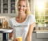 Eine lächelnde Frau in einer modernen, hellen Küche, die ein Glas frisch gepressten Saft in der Hand hält. Sie trägt ein weißes T-Shirt mit dem Aufdruck „Plant Power – No Limits.com“, während neben ihr ein Slow Juicer steht, umgeben von frischem Obst und Gemüse.