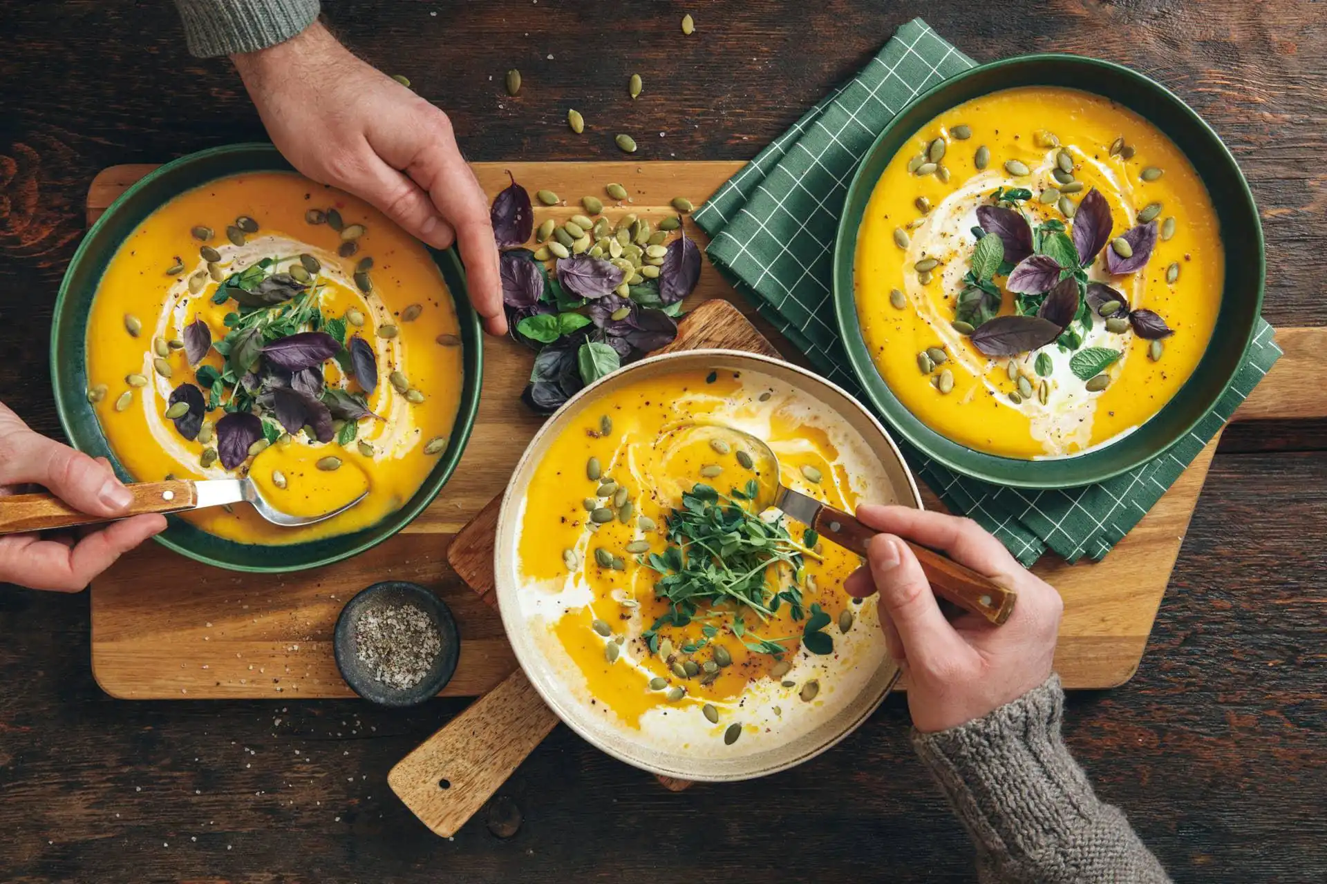 Drei Schüsseln cremige Kürbissuppe mit Kokosmilch, frischen Kräutern und Kürbiskernen auf einem gedeckten Holztisch.
