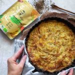 Goldbraun gebackenes Kartoffelgratin in einer runden Backform, dekoriert mit Bio Pommes de Terre Food Finisher, der in einer Verpackung daneben liegt.
