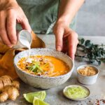Ein Teller mit cremiger asiatischer Kürbissuppe, garniert mit frischen Kräutern und Kürbiskernen, auf einem rustikalen Holztisch.