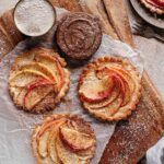 Drei Apfeltörtchen mit fein geschnittenen Apfelscheiben, die mit Zimt bestäubt sind, auf einem Holzbrett angerichtet. Daneben befinden sich eine Schale mit Schokocreme und ein Sieb mit Puderzucker.