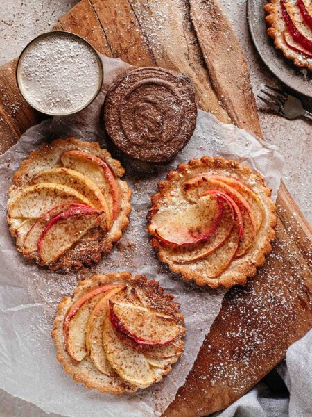 Drei Apfeltörtchen mit fein geschnittenen Apfelscheiben, die mit Zimt bestäubt sind, auf einem Holzbrett angerichtet. Daneben befinden sich eine Schale mit Schokocreme und ein Sieb mit Puderzucker.