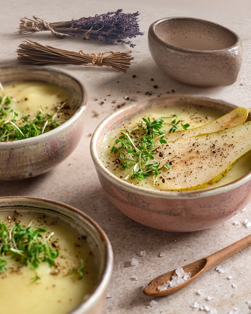 Geröstete Pastinaken-Birnen-Suppe mit frischer Kresse, Birnenscheiben und Pfeffer, serviert in rustikalen Keramikschalen, dekoriert mit Lavendel und Meersalz.