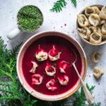 Eine Schale mit Rote-Bete-Suppe und Tortellini, umgeben von einer Schale (Bio Braten-Magic Food Finisher) und Teigtaschen, eingerahmt von grünen Tannenzweigen.