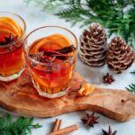 Zwei Gläser mit heißem Gewürztee oder Punsch, dekoriert mit Orangenscheiben, Zimtstangen und Sternanis, stehen auf einem rustikalen Holzbrett. Im Hintergrund sind Tannenzweige und dekorative Tannenzapfen zu sehen, die eine gemütliche Winteratmosphäre schaffen.