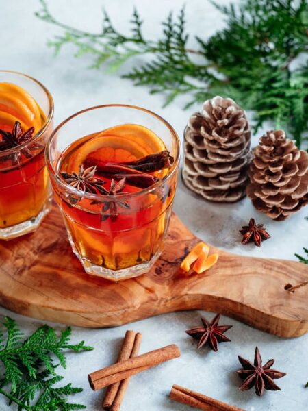 Zwei Gläser mit heißem Gewürztee oder Punsch, dekoriert mit Orangenscheiben, Zimtstangen und Sternanis, stehen auf einem rustikalen Holzbrett. Im Hintergrund sind Tannenzweige und dekorative Tannenzapfen zu sehen, die eine gemütliche Winteratmosphäre schaffen.