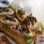 Crostini aus Baguettescheiben, belegt mit Kräuter-Frischkäse und sautierten Pilzen, dekoriert mit frischen Kräutern auf einem Holzbrett. Ein mediterraner Snack in einladender Präsentation.