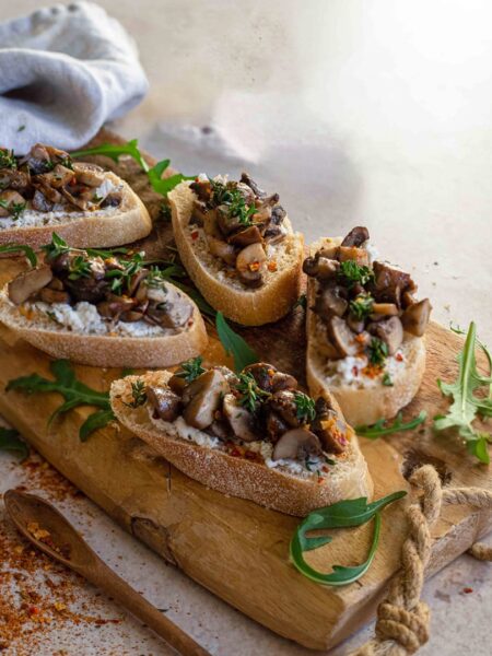 Crostini aus Baguettescheiben, belegt mit Kräuter-Frischkäse und sautierten Pilzen, dekoriert mit frischen Kräutern auf einem Holzbrett. Ein mediterraner Snack in einladender Präsentation.