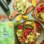 Mediterraner Nudelsalat mit roher Zucchini, Kichererbsen, Tomaten und kurzen Nudeln. Frisch serviert mit Zitronenspalten und dünn geschnittenen Zucchinistreifen.