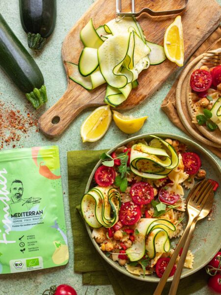 Mediterraner Nudelsalat mit roher Zucchini, Kichererbsen, Tomaten und kurzen Nudeln. Frisch serviert mit Zitronenspalten und dünn geschnittenen Zucchinistreifen.