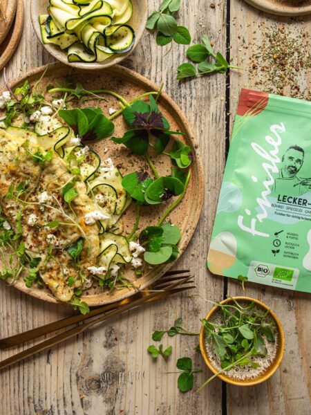 Ein Zucchini-Ziegenkäse-Omelett auf einem rustikalen Teller, dekoriert mit Kräutern und Zucchinistreifen, daneben frisches Brot und die grüne Verpackung des Bio Lecker-Ei Food Finishers.