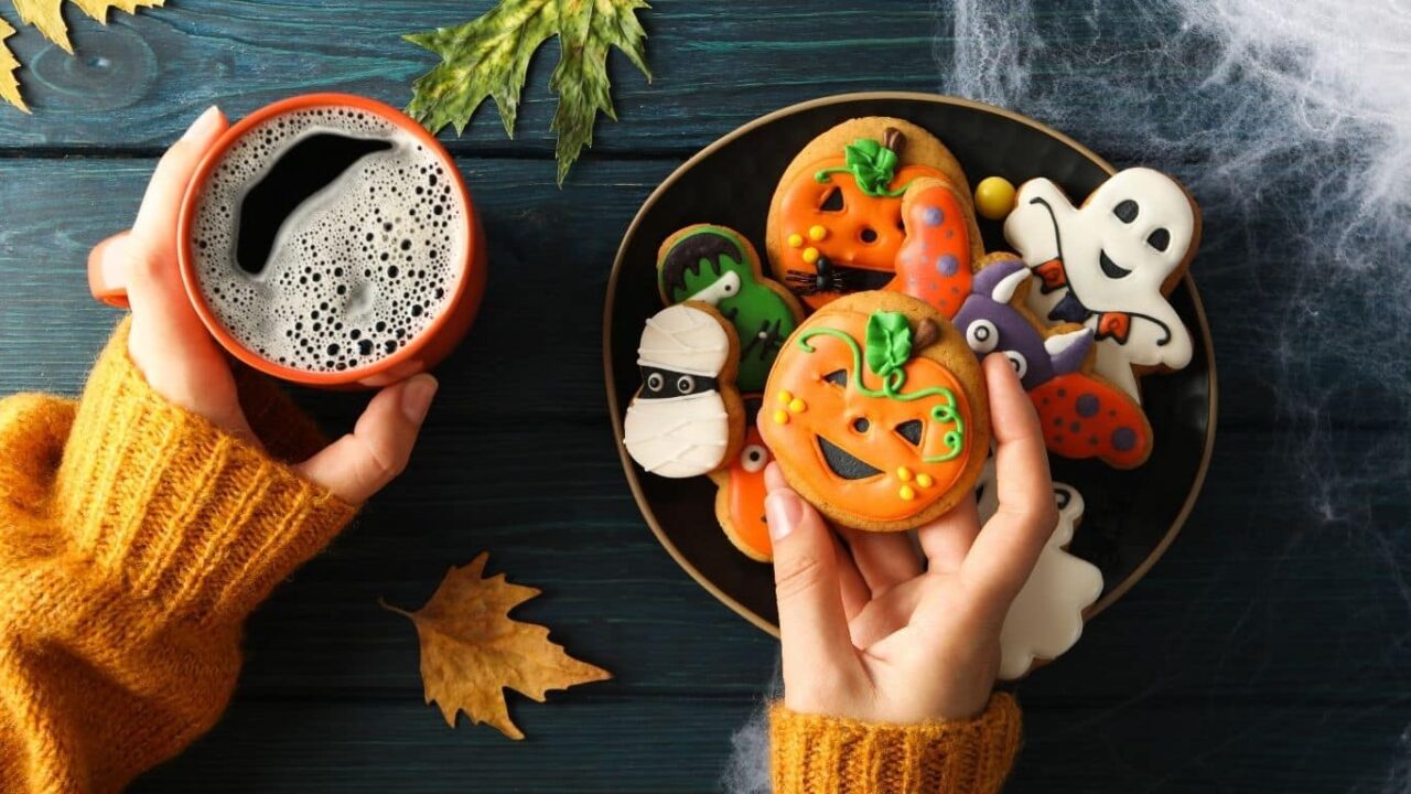 Frau hält eine Tasse Kaffee in der einen und bunte Halloween Cookies in der anderen Hand. Im Hintergrund herbstliche Deko mit Kürbis, Blättern und Food Finisher Spices. Perfekt für herbstliche Backideen und Halloween-Rezepte mit Bio-Gewürzen. 100 % Bio, vegan, ohne Zusatzstoffe.