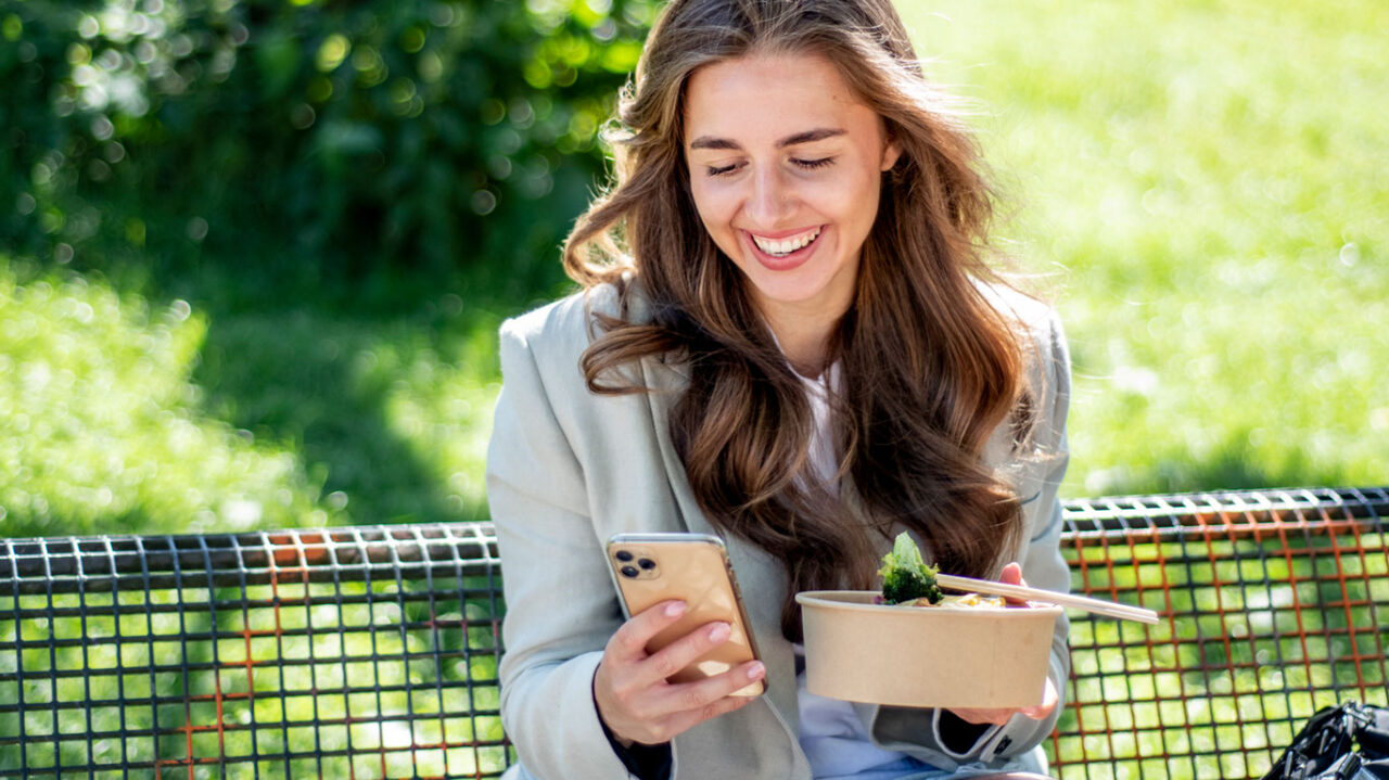 Eine Frau sitzt auf einer Parkbank, genießt eine Mahlzeit und schaut gleichzeitig auf ihr Smartphone. Umgeben von weicher natürlicher Beleuchtung und der ruhigen Atmosphäre des Parks, zeigt die Szene die moderne Ablenkung durch digitale Geräte, selbst während des Essens.