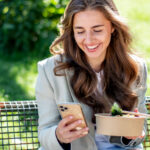 Eine Frau sitzt auf einer Parkbank, genießt eine Mahlzeit und schaut gleichzeitig auf ihr Smartphone. Umgeben von weicher natürlicher Beleuchtung und der ruhigen Atmosphäre des Parks, zeigt die Szene die moderne Ablenkung durch digitale Geräte, selbst während des Essens.