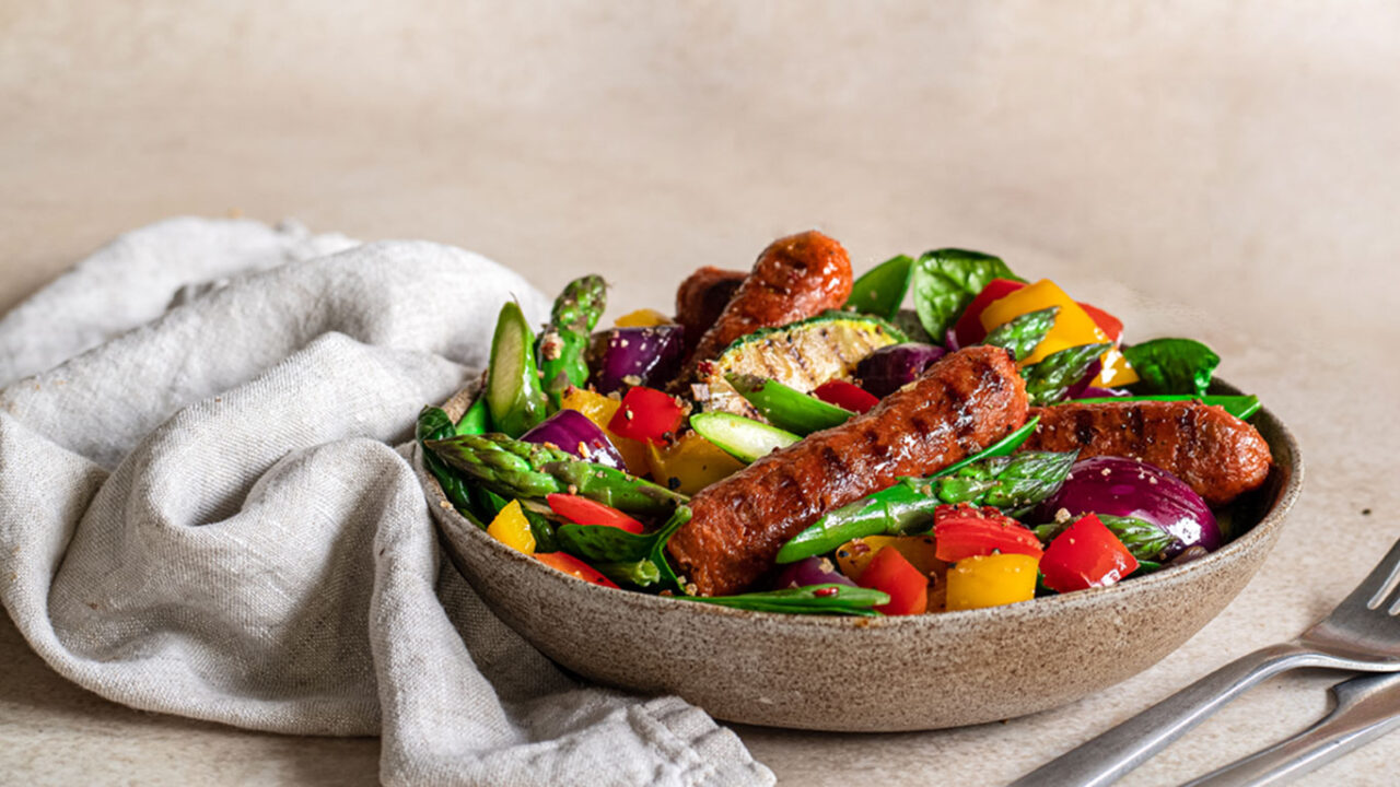 Ein Teller mit veganer Wurst, serviert zusammen mit frischem Gemüse wie Paprika, Zucchini und Tomaten. Die farbenfrohe Mahlzeit präsentiert eine ausgewogene und nachhaltige Alternative zu Fleisch, ideal für eine gesunde und pflanzliche Ernährung