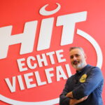 Luis Dias in einem blauen Hemd steht vor einer großen roten Wand mit dem weißen Logo von HIT-Märkten, das den Schriftzug "HIT Echte Vielfalt" zeigt. Er lächelt freundlich und hat die Arme locker verschränkt.