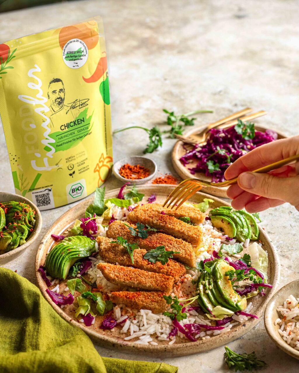 Lunch-Bowl mit Reis, Avocado, Rotkohl und knusprigen pflanzlichen Hähnchenstreifen, verfeinert mit der Gewürzmischung "Bio Chicken Food Finisher" von Food Finisher Spices. Im Hintergrund ist die gelbe Produktverpackung zu sehen, während eine Hand mit einer goldenen Gabel nach den Hähnchenstreifen greift.