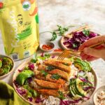 Lunch-Bowl mit Reis, Avocado, Rotkohl und knusprigen pflanzlichen Hähnchenstreifen, verfeinert mit der Gewürzmischung "Bio Chicken Food Finisher" von Food Finisher Spices. Im Hintergrund ist die gelbe Produktverpackung zu sehen, während eine Hand mit einer goldenen Gabel nach den Hähnchenstreifen greift.