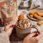 Eine Person hält eine Tasse mit heißer Schokolade, die mit Schlagsahne, Schokoladenraspeln und einem Keks dekoriert ist. Im Hintergrund sind Kekse auf einem Teller und eine Verpackung für Kakao-Pulver zu sehen.