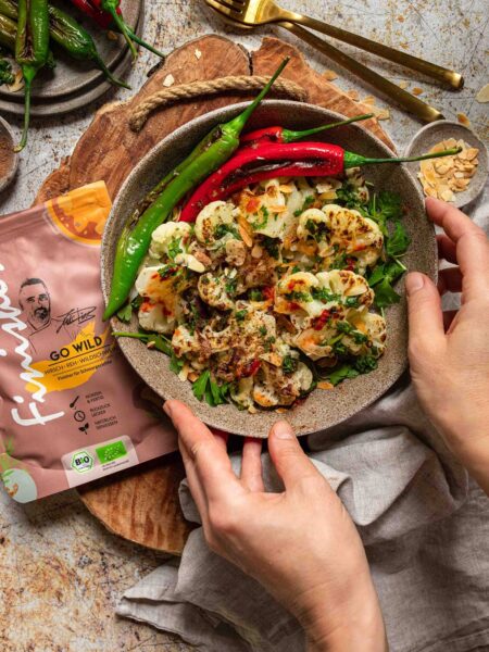 Ein Teller mit geröstetem Blumenkohl, dekoriert mit roten und grünen Peperoni. Daneben liegt eine Packung des Bio Go Wild Food Finisher von Food Finisher Spices.