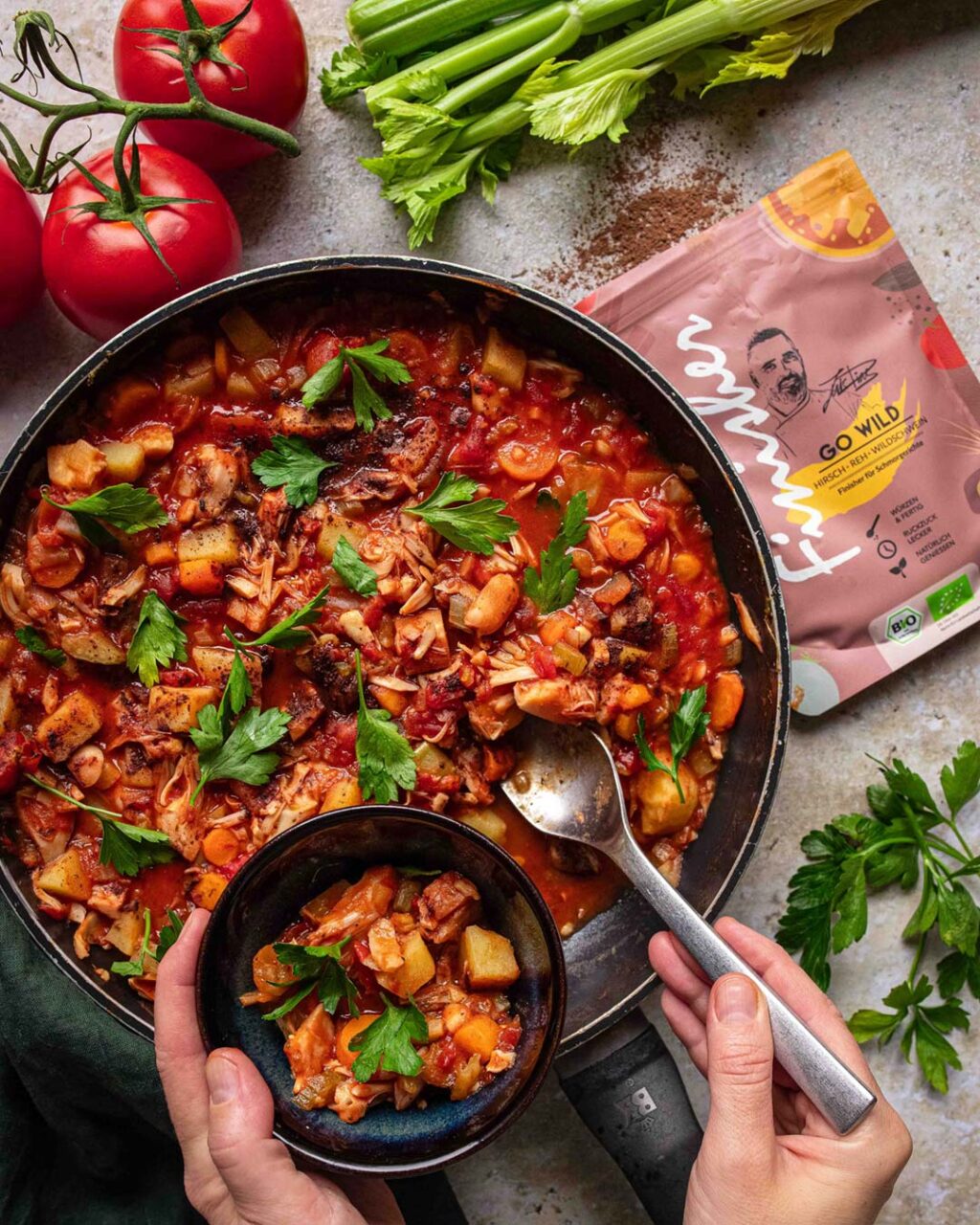 Jackfruit- und Kartoffeleintopf in einer Pfanne mit frischen Kräutern, serviert in einer kleinen Schüssel. Im Hintergrund liegen frische Zutaten wie Tomaten, Sellerie und Petersilie sowie eine Verpackung des Bio Wild Food Finisher.