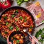 Jackfruit- und Kartoffeleintopf in einer Pfanne mit frischen Kräutern, serviert in einer kleinen Schüssel. Im Hintergrund liegen frische Zutaten wie Tomaten, Sellerie und Petersilie sowie eine Verpackung des Bio Wild Food Finisher.