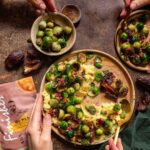 Hände greifen nach einem Gericht aus geröstetem Rosenkohl, Datteln und cremigem Püree, gewürzt mit dem Bio Go Wild Food Finisher.