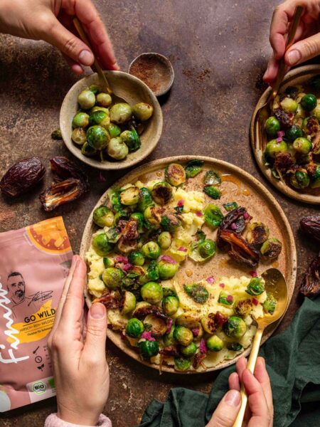 Hände greifen nach einem Gericht aus geröstetem Rosenkohl, Datteln und cremigem Püree, gewürzt mit dem Bio Go Wild Food Finisher.